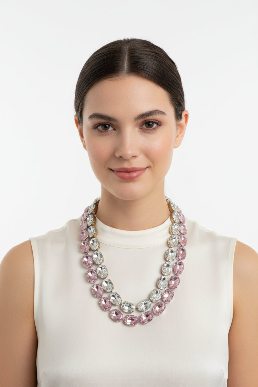 A close-up of a hand holding a multi-strand necklace made of faceted oval rhinestones in pink, clear, and silver tones against a plain light gray background.