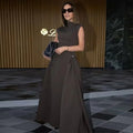 Woman in a brown dress holding a handbag in an indoor setting with a gold logo in the corner.