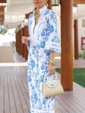 Woman in a blue and white floral dress holding a beige handbag on a wooden deck.