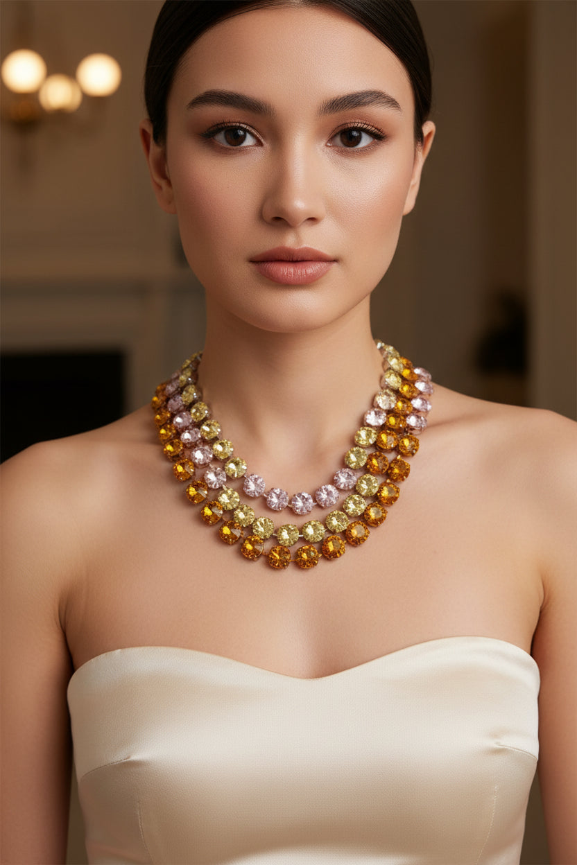 A hand holding a strand of faceted rhinestone beads in multiple colors, including amber orange, pale yellow, lavender pink, and light blue, on a plain light gray background.