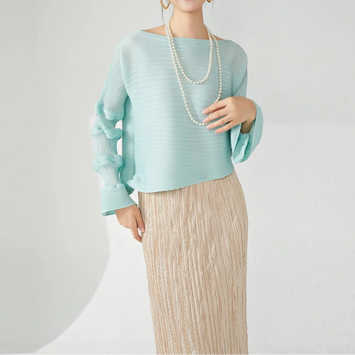 A woman modeling a light mint green long-sleeve top with a boat neckline and pleated texture, paired with a beige pleated skirt and a double-strand pearl necklace, against a clean white studio background.