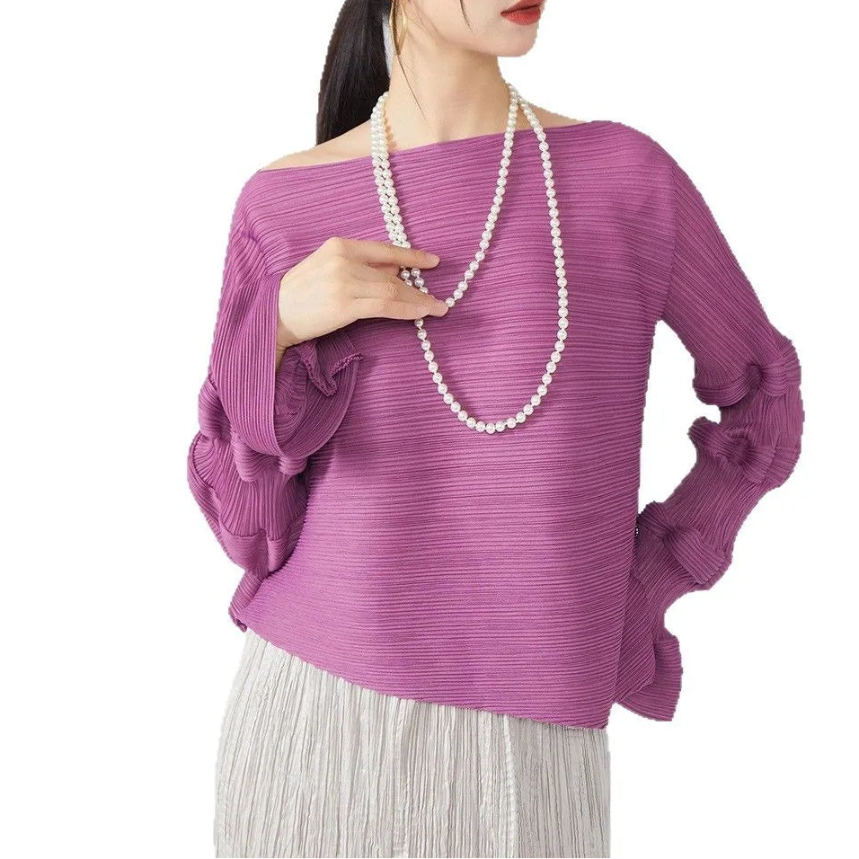 A woman wearing a textured off-the-shoulder purple knit top with long ruffled sleeves, paired with a long white pearl necklace and light gray pleated bottoms, set against a plain white background.