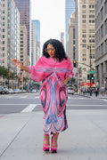 Person wearing a vibrant pink and blue patterned dress standing outdoors with greenery in the background.