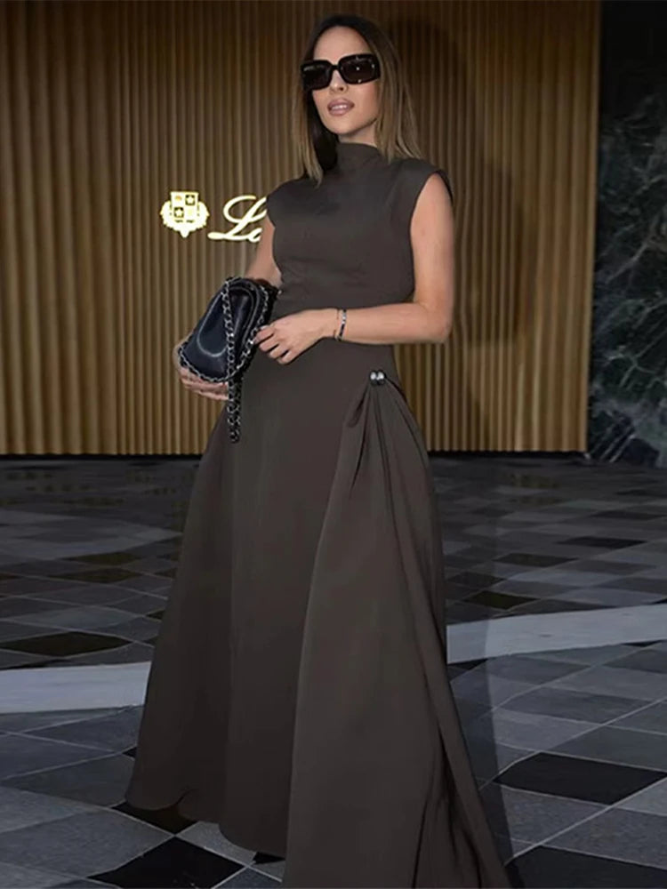 Woman in a brown dress holding a handbag in an indoor setting with a gold logo in the corner.