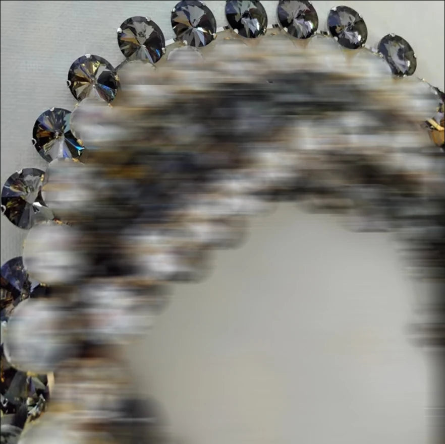 A close-up of a circular product with a border of faceted dark gray to black rhinestones, shown against a light gray background. The center of the circle is heavily blurred or obscured.