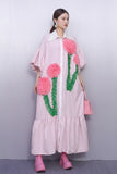 Woman wearing a pink dress with floral designs and holding a pink handbag against a gray background