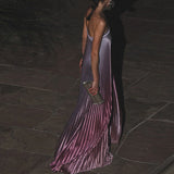 Woman in a long, pleated dress holding a clutch on a dark stone floor.