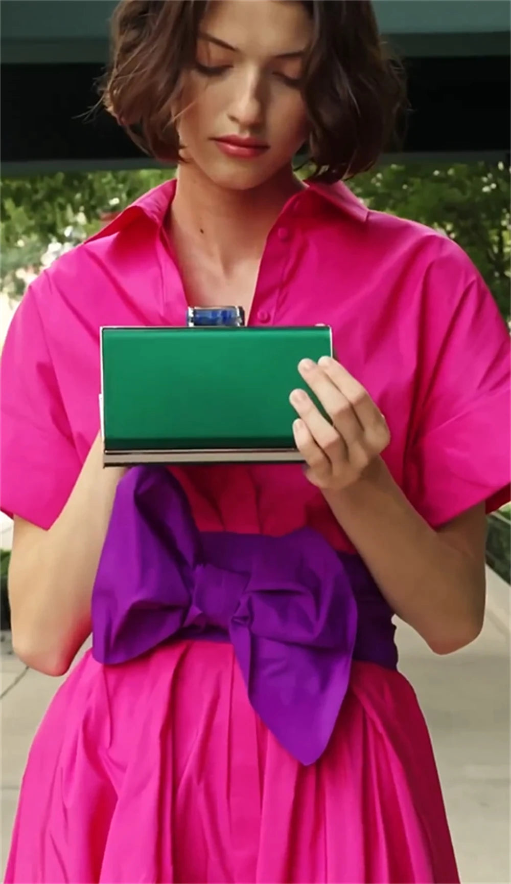 Woman in a bright pink dress with a purple bow holding a green clutch.