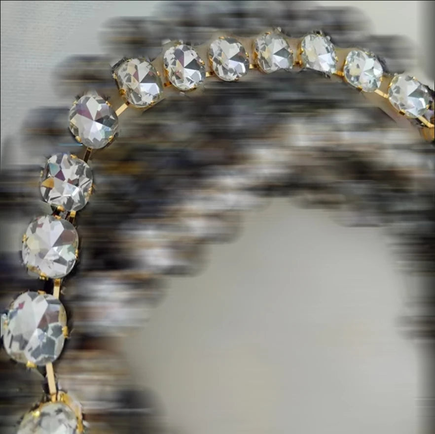 A close-up of a gold-toned jewelry piece, likely a bracelet or necklace, adorned with large faceted clear rhinestones arranged in an arch. The background is heavily blurred and out of focus.