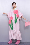 Woman wearing a pink dress with floral designs and holding a matching handbag against a plain background