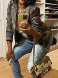 Person wearing a camouflage jacket and blue jeans, holding a gold glittery handbag.