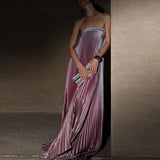 Woman in a strapless pleated dress holding a clutch against a neutral wall.
