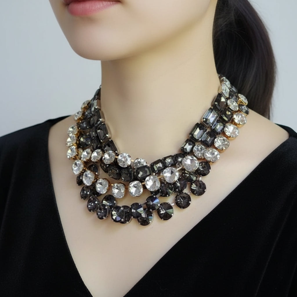Close-up of a multi-strand crystal necklace featuring round, oval, and rectangular faceted rhinestones in clear, silver, and dark smoky gray tones, set in gold-toned metal on a light beige background.