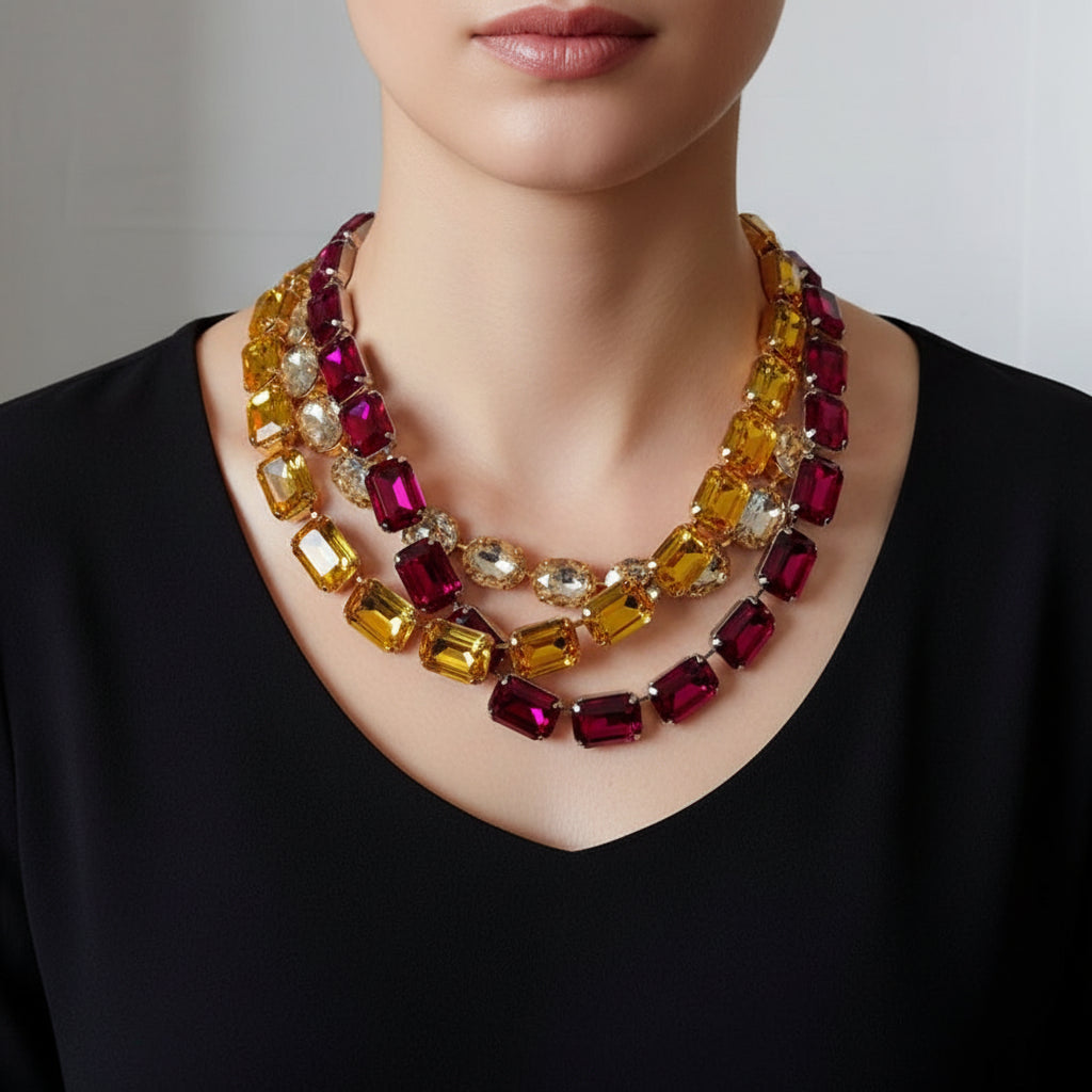 A multi-strand statement necklace made of large rectangular and round faceted rhinestones in yellow, deep magenta, and clear, displayed on a textured beige jewelry bust.