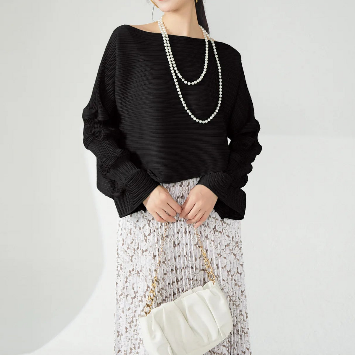 A woman modeling a black ribbed long-sleeve top with a wide boat neckline, paired with a white patterned skirt and a double-strand pearl necklace, holding a small white handbag against a plain light gray background.