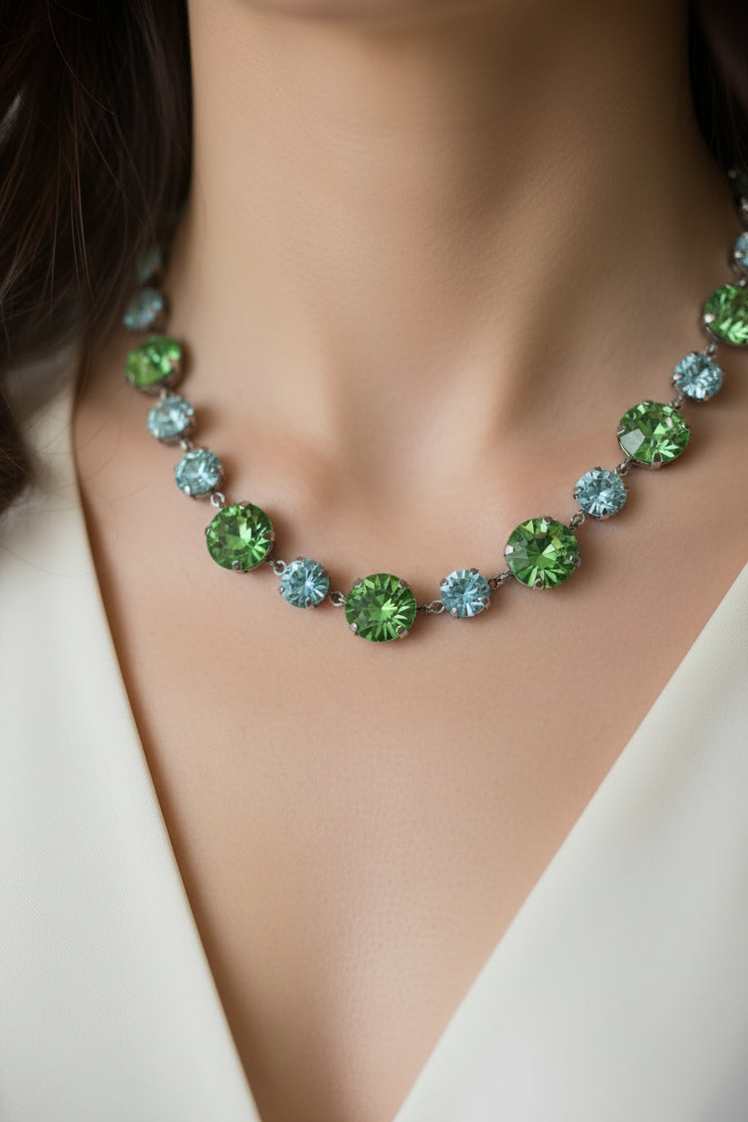 A beaded necklace and matching bracelet laid out on a light beige textured background. The jewelry features faceted green and light blue rhinestones set in silver-tone metal, with a lobster clasp closure.