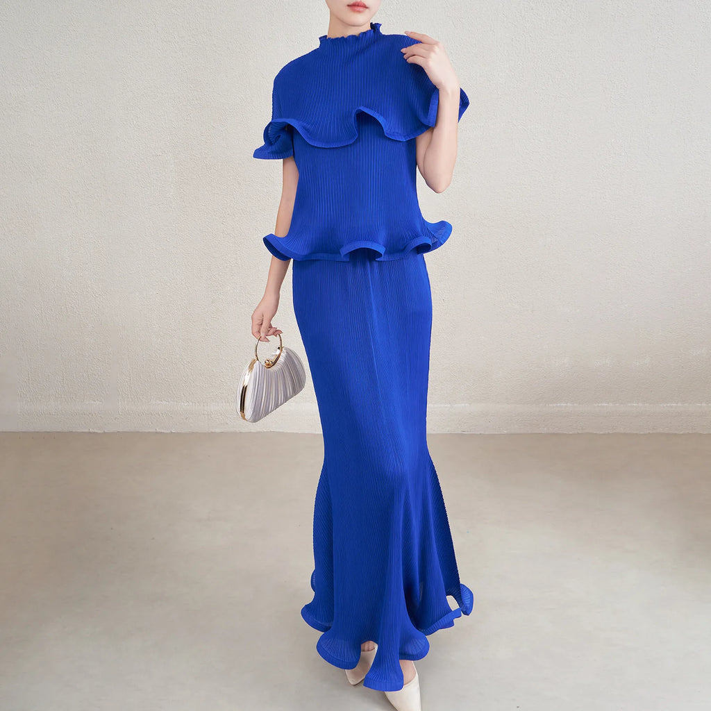 Woman wearing a blue dress with ruffled top and skirt, holding a silver handbag against a plain background.