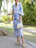 Woman in a blue floral dress with a white kimono-style jacket, standing outdoors with greenery in the background.