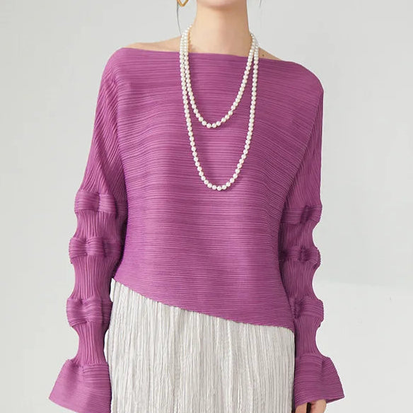 A woman wearing a textured off-the-shoulder knit sweater in a dusty purple/plum color, styled with a double-strand pearl necklace and a pleated light gray skirt, against a plain light gray background.