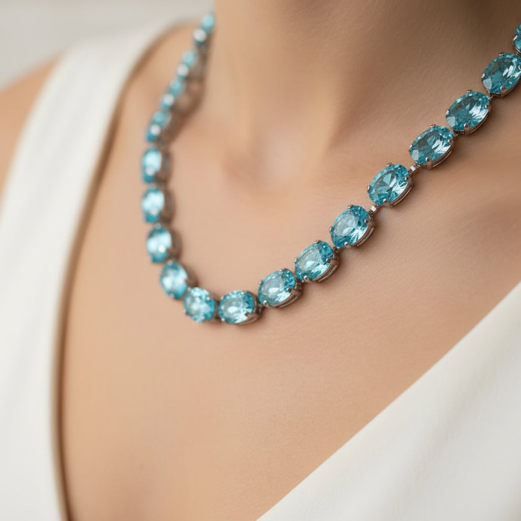 A silver-toned tennis bracelet featuring a row of oval-cut, light blue gemstones, displayed on a neutral light beige background.