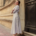 Woman in a light gray dress with floral embroidery leaning against a stone wall.