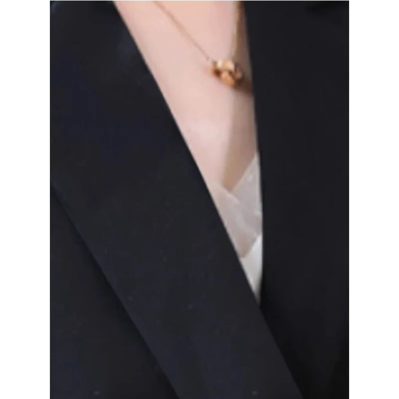 A close-up of a person wearing a delicate gold necklace with a small round pendant, styled with a black blazer and a white top.