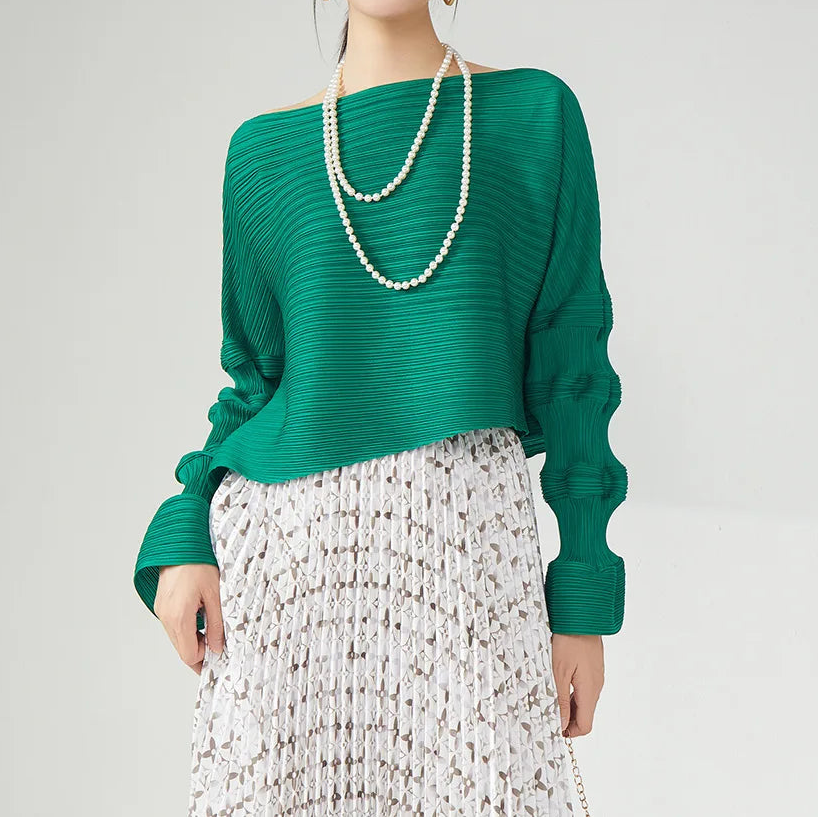 A woman modeling a green long-sleeve knit top with a wide off-shoulder neckline, paired with a textured white skirt and layered pearl necklaces, against a light gray studio background.
