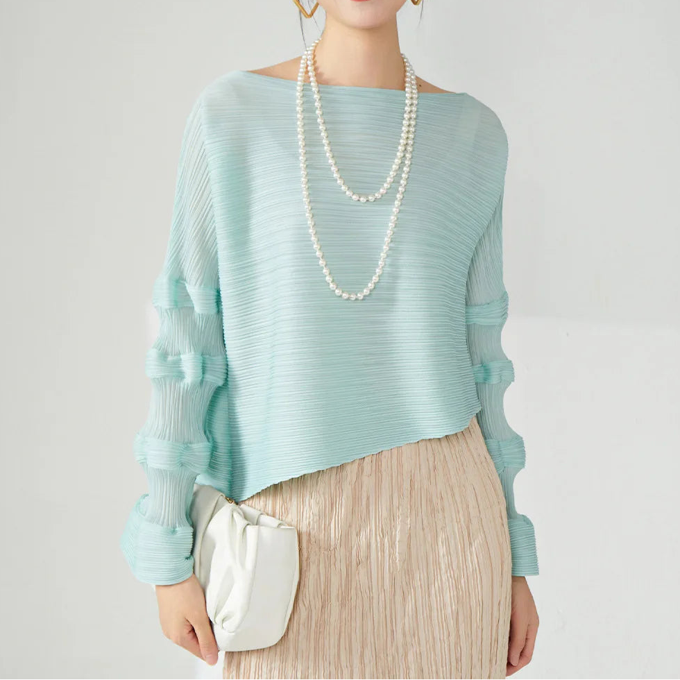 A woman models a light mint green knit top with sheer ruffled sleeves, paired with a beige pleated skirt and accessorized with a double-strand pearl necklace and a small white crossbody bag, against a plain light gray background.