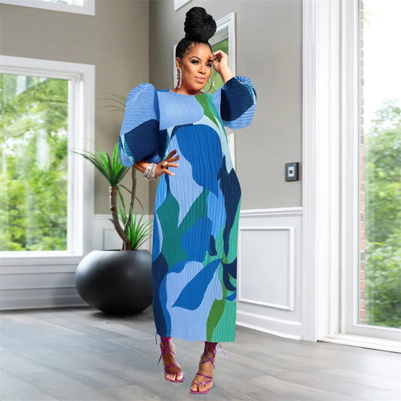 Woman in a blue and green dress standing in a room with large windows and plants.