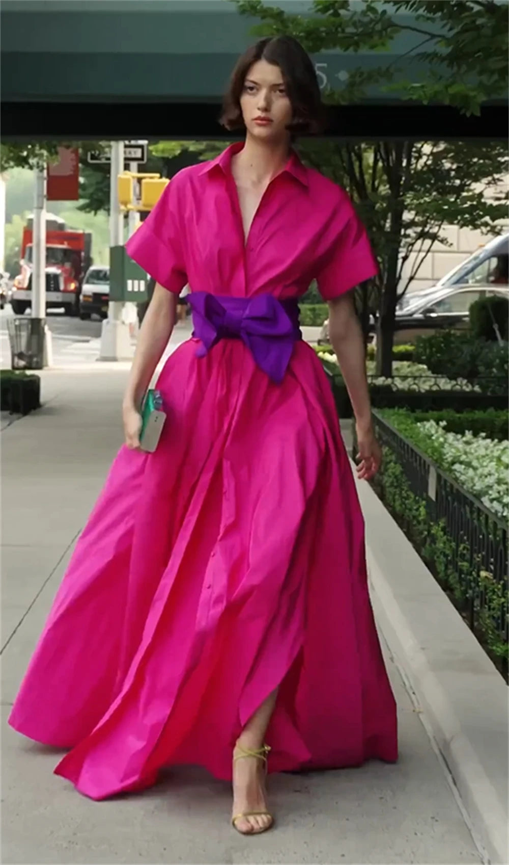 Woman in a bright pink dress with a purple belt walking on a sidewalk.