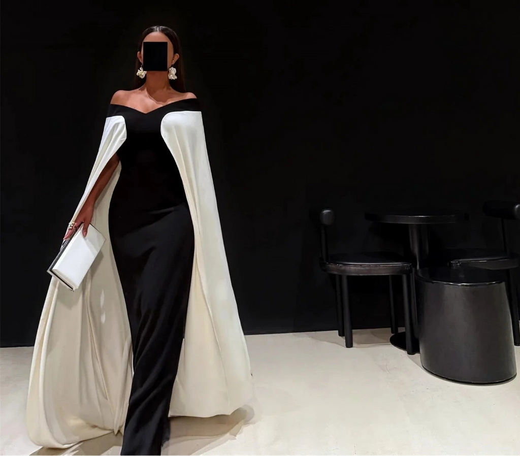 Woman in a black and white dress with a cape holding a clutch against a black background.