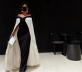 Woman in a black and white dress with a cape holding a clutch against a black background.