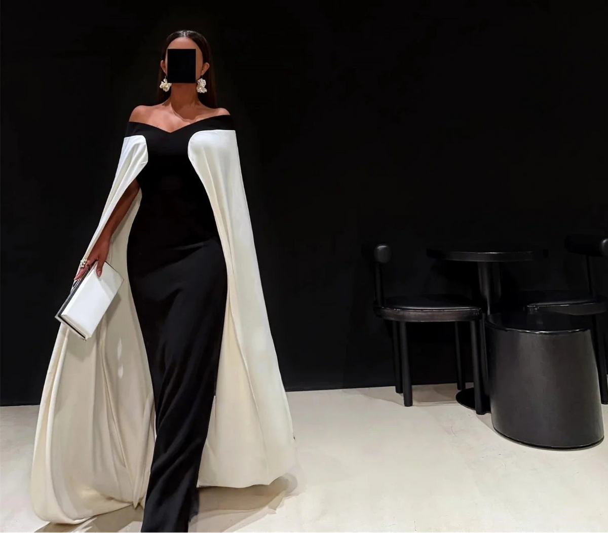 Woman in a black and white dress with a cape holding a clutch against a black background.