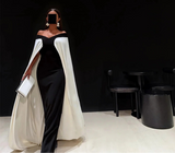 Woman in a black and white dress with a cape holding a clutch against a black background.