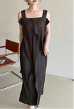 Woman wearing a dark brown pleated dress standing in a minimalistic room with a chair and light-colored floor.