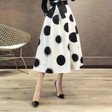 Person wearing a black top and white skirt with black polka dots in a minimalistic room.