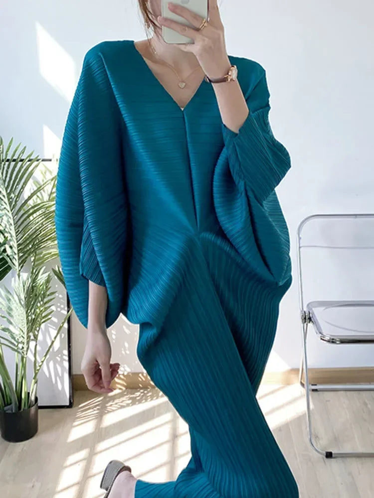 Person wearing a teal ribbed sweater and pants in a bright room with plants and a chair.